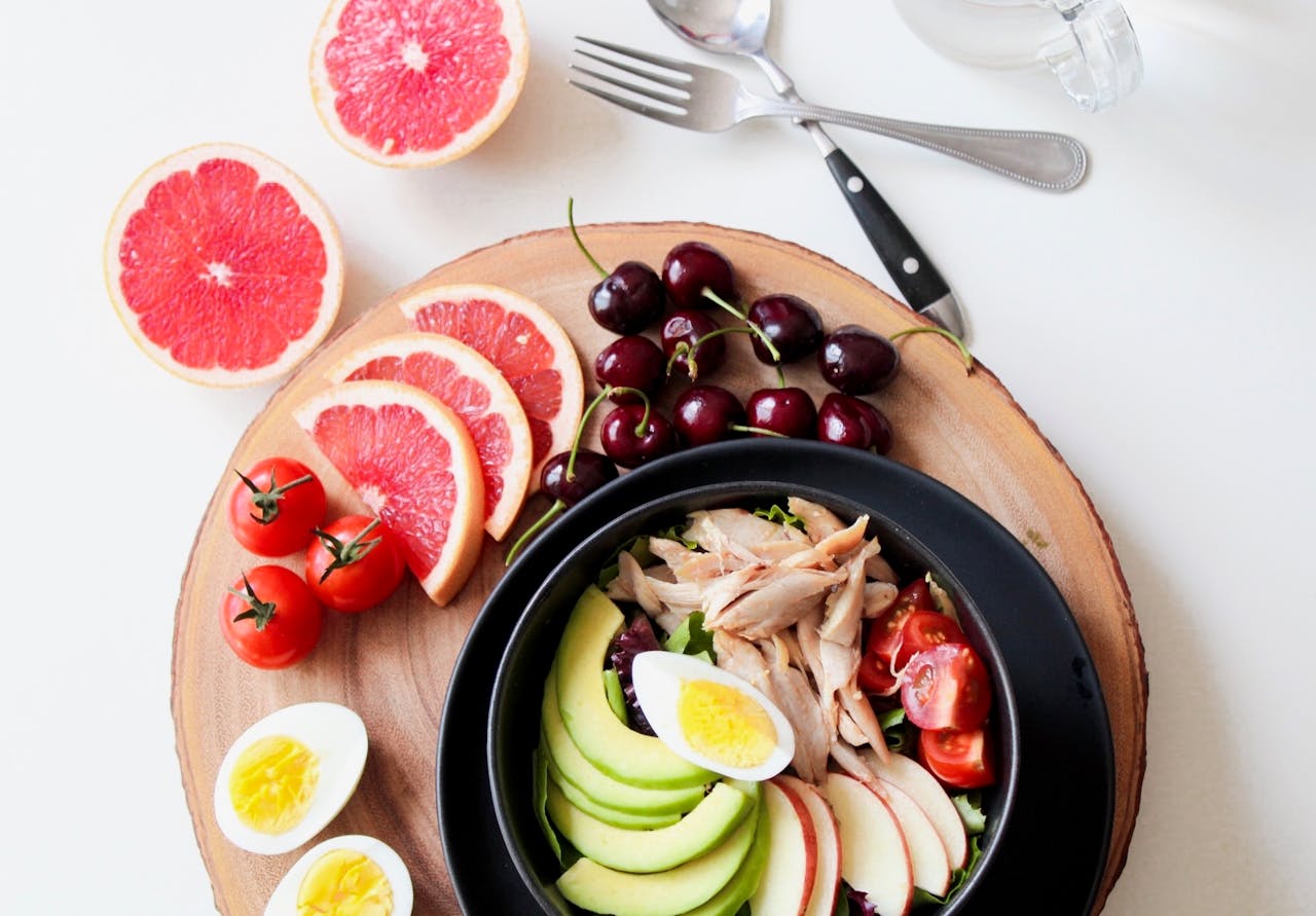 services-04 A vibrant breakfast flat lay featuring a mix of fruits, avocado, eggs, tomatoes, and sliced meats on a wooden platter.